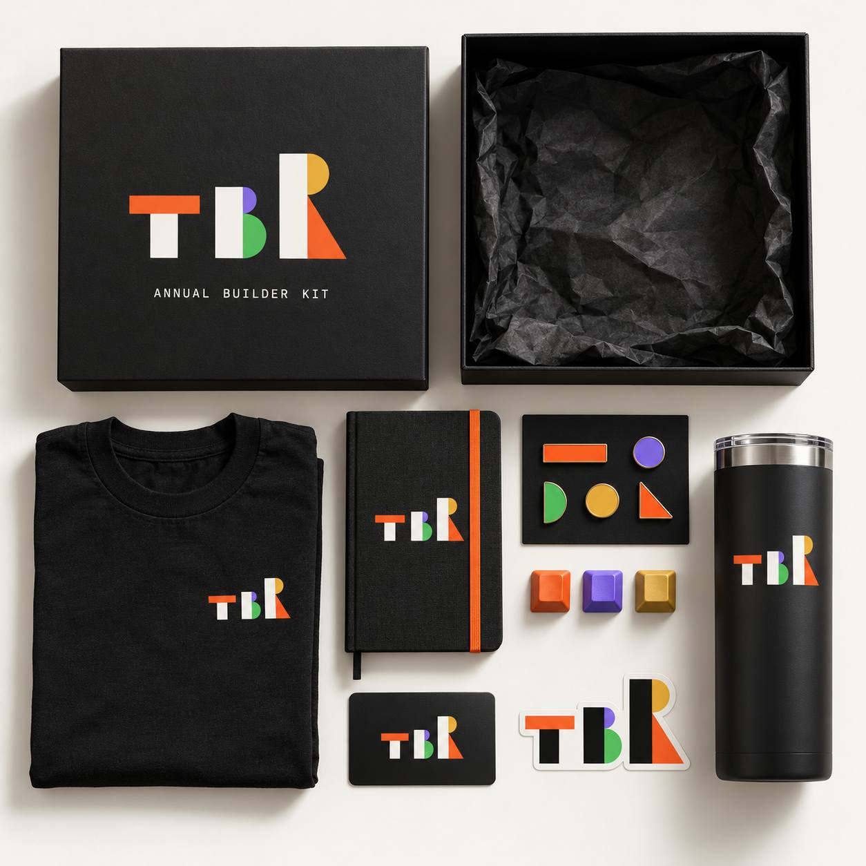 Kotak hitam Annual Builder Kit terbuka, dikelilingi kaos, buku catatan, pin, keycap, kartu member, stiker, dan tumbler berlogo TBR.