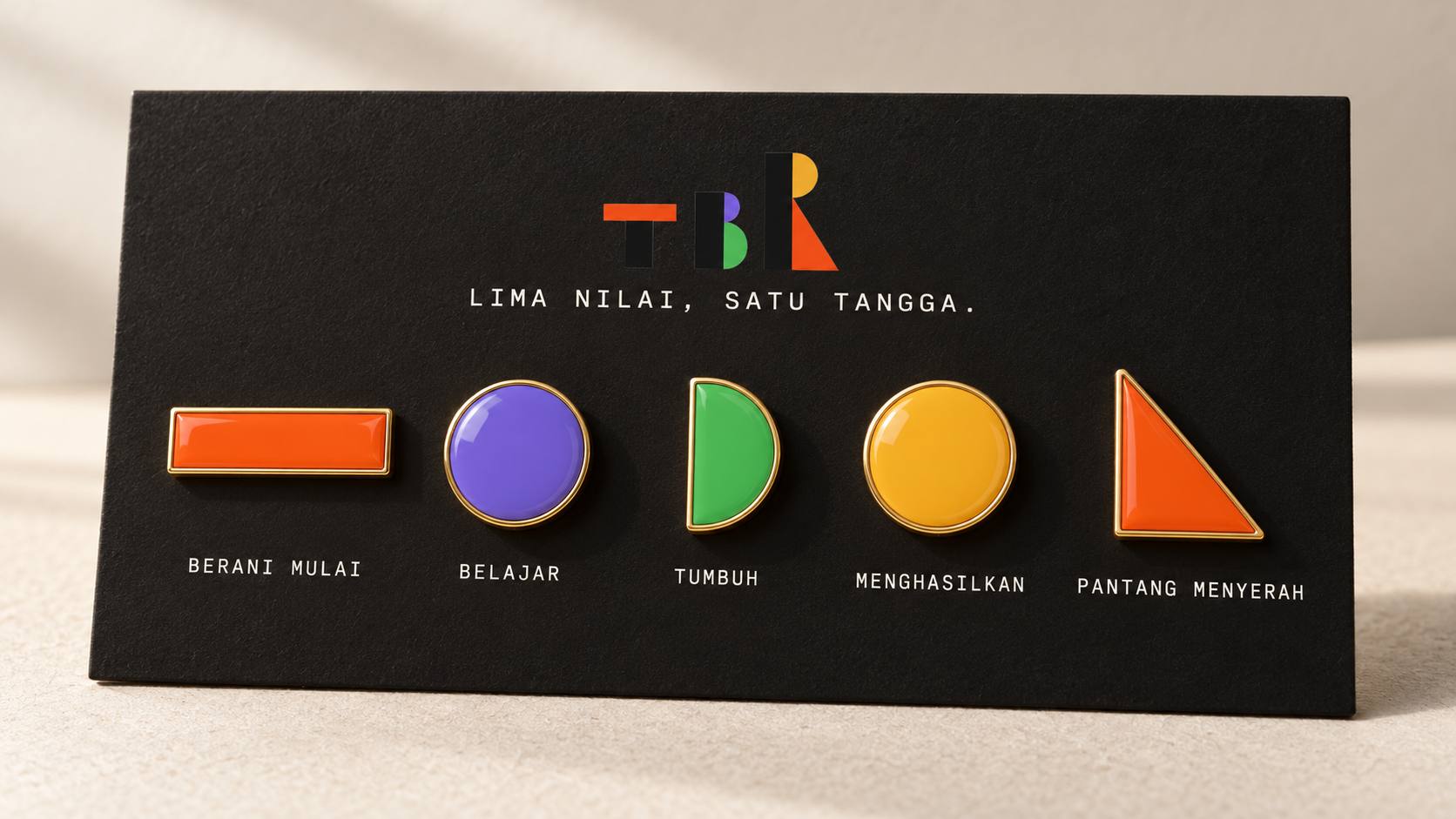 Lima pin enamel berbentuk potongan logo TBR di kartu hitam, satu untuk tiap nilai: berani mulai, belajar, tumbuh, menghasilkan, pantang menyerah.