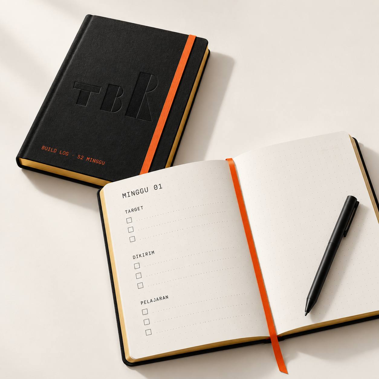 Buku catatan BUILD LOG 52 MINGGU bersampul linen hitam, terbuka di halaman MINGGU 01.